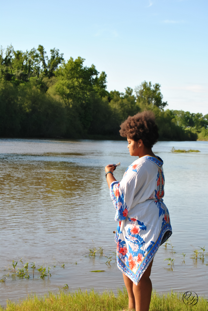 Jamelah standing on a river bank holding a small smoking bundle of sage in her left hand and a piece of smoking cedar wood in her right by her leg. Facing the river (left) and has a calm/peaceful expression with a cursive 'J T L' watermark in the bottom right corner