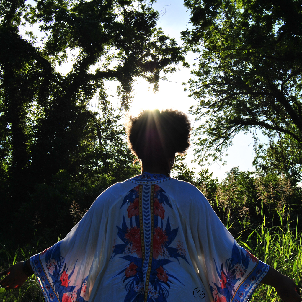 Jamelah facing away form the camera with arms out reached and the sun illumenating her afro and clothing in a outdoor setting and a JTL water mark on the lower right side