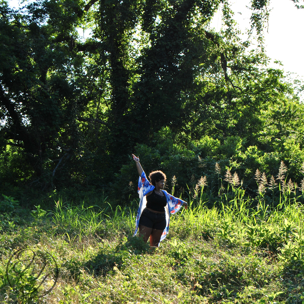 Jamelah standing in a field with arms raised as if dancing amongst the flora with a cursive JTL logo in the bottom left corner