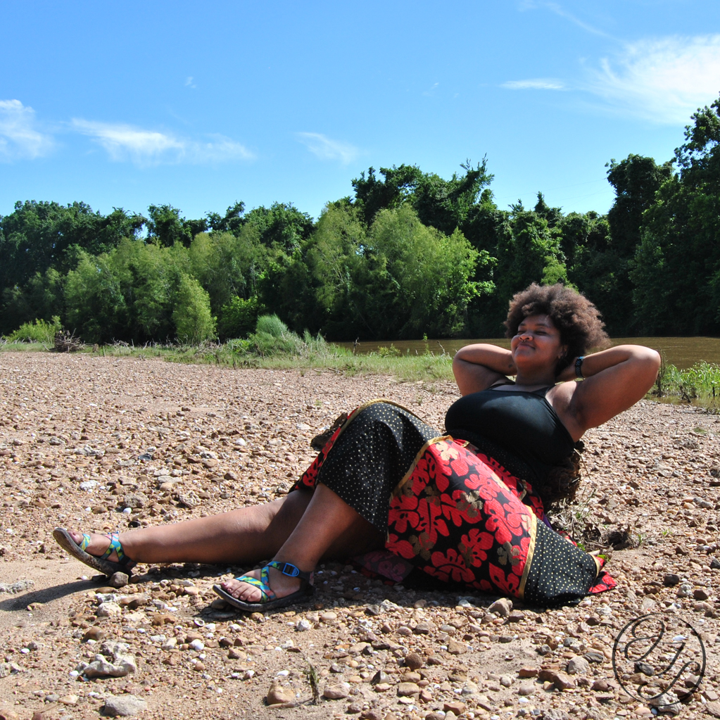 Jamelah on a river bank with her arms propped up resting and a JTL water mark in lower right corner