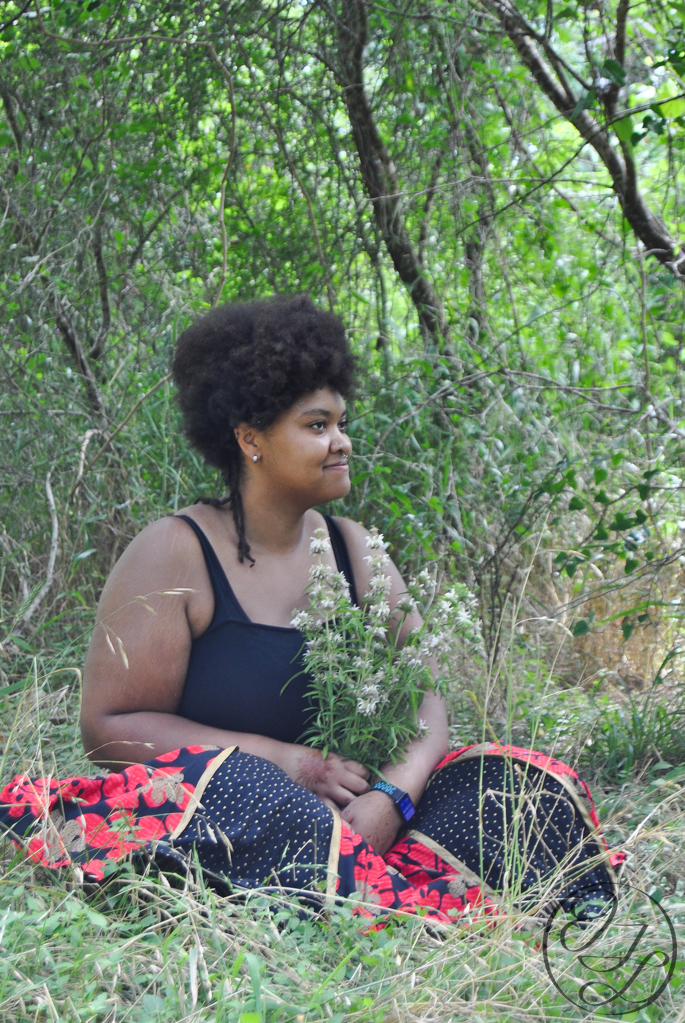 Jamelah sitting in some grass in a shady spot with a bouque of bee balm in her lap, she is looking off to the side with calm smile and look on her face, a cursive JTL logo is in the bottom right corner