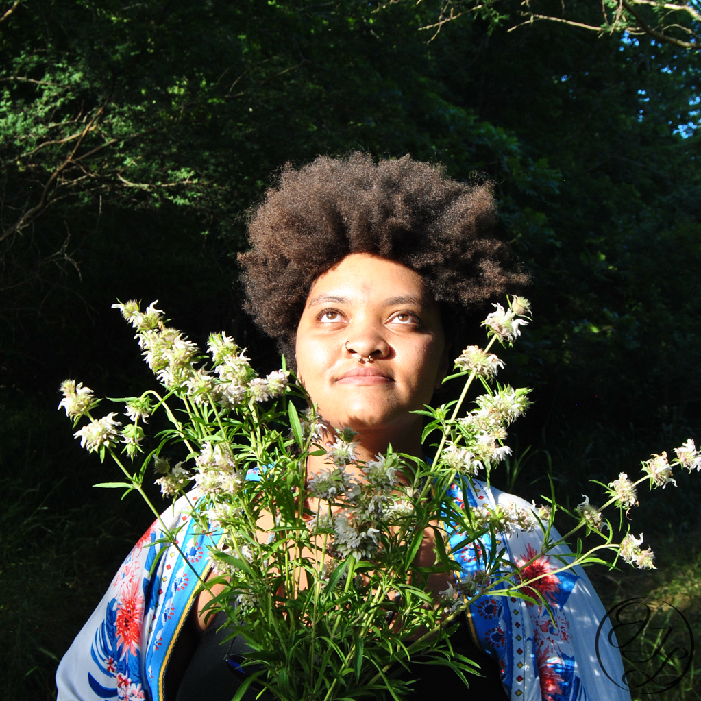 Jamelah gazing up while the sun beams on her face from the left a bouquet of lemon beebalm framing her face and a JTL logo in the lower right corner