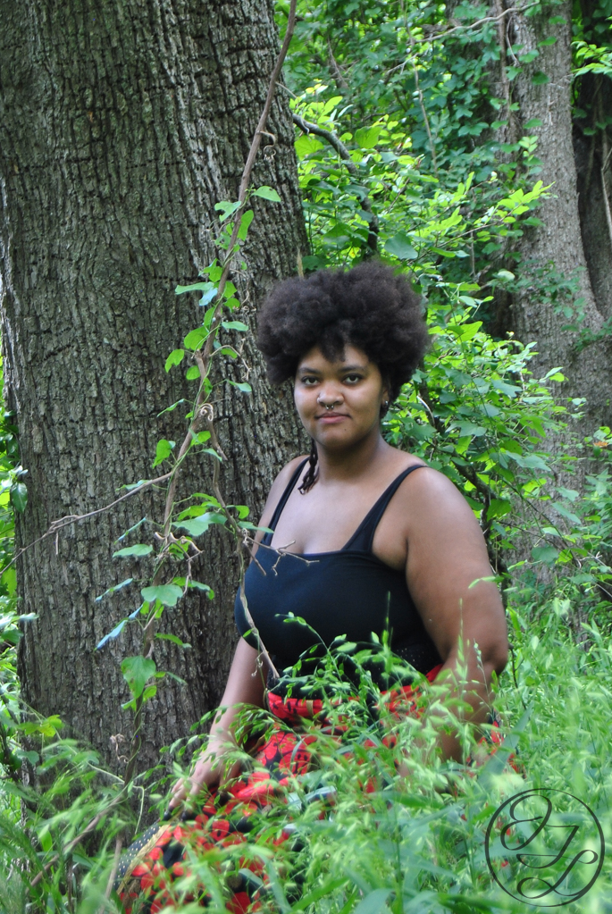 Jamelah standing in front of a cottonwood tree behind some tall grass and a vine in front of her, she is looking directly at the camera, there is a circular watermark that reads 'JTL' in cursive on the bottom right corner.