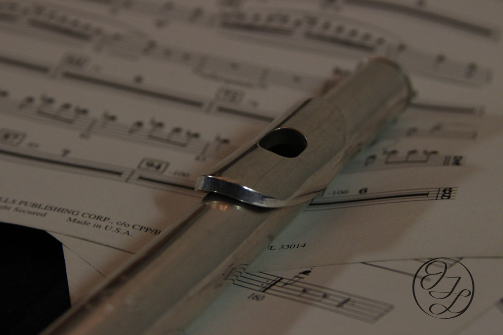 A silver flute laying on some sheet music with a cursive JTL with a circle around it in the bottom right corner