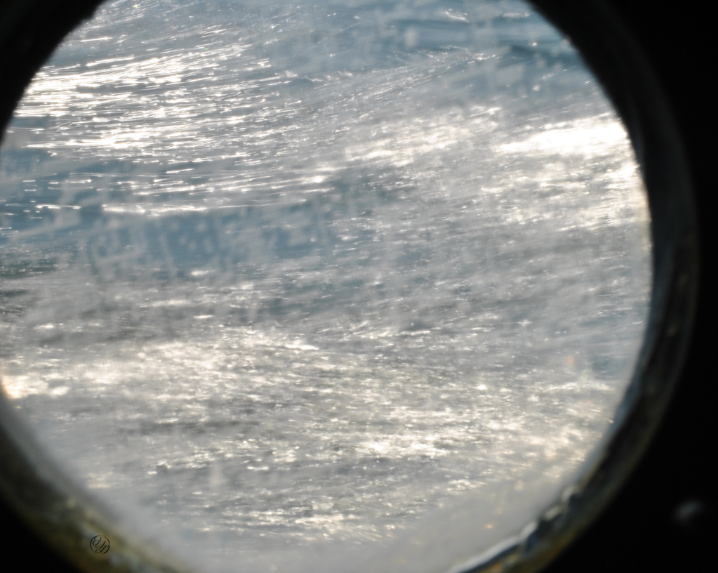 Crisp clear surf through a scratched port hole and a hidden cursive JTL, with a circle around it, logo