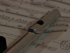 A silver flute laying on some sheet music with a cursive JTL with a circle around it in the bottom right corner