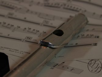 27 Wk w_ WM sq. A silver flute laying on some sheet music with a cursive JTL with a circle around it in the bottom right corner