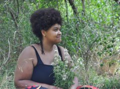 Jamelah sitting in some grass in a shady spot with a bouque of bee balm in her lap, she is looking off to the side with calm smile and look on her face, a cursive JTL logo is in the bottom right corner