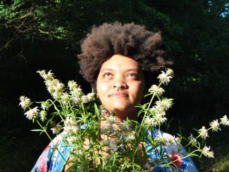 Jamelah gazing up while the sun beams on her face from the left a bouquet of lemon beebalm framing her face and a JTL logo in the lower right corner