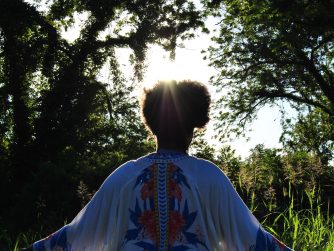 Jamelah facing away form the camera with arms out reached and the sun illumenating her afro and clothing in a outdoor setting and a JTL water mark on the lower right side