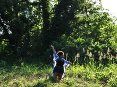 Jamelah standing in a field with arms raised as if dancing amongst the flora with a cursive JTL logo in the bottom left corner