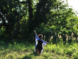 Jamelah standing in a field with arms raised as if dancing amongst the flora with a cursive JTL logo in the bottom left corner