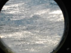 Crisp clear surf through a scratched port hole and a hidden cursive JTL, with a circle around it, logo