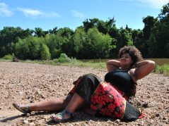 Jamelah on a river bank with her arms propped up resting and a JTL water mark in lower right corner