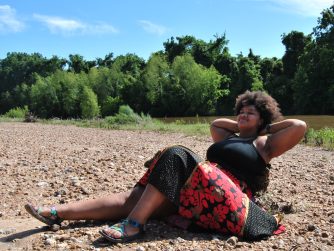 Jamelah on a river bank with her arms propped up resting and a JTL water mark in lower right corner