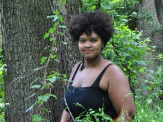 Jamelah standing in front of a cottonwood tree behind some tall grass and a vine in front of her, she is looking directly at the camera, there is a circular watermark that reads 'JTL' in cursive on the bottom right corner.