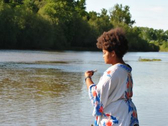 Jamelah standing on a river bank holding a small smoking bundle of sage in her left hand and a piece of smoking cedar wood in her right by her leg. Facing the river (left) and has a calm/peaceful expression with a cursive 'J T L' watermark in the bottom right corner